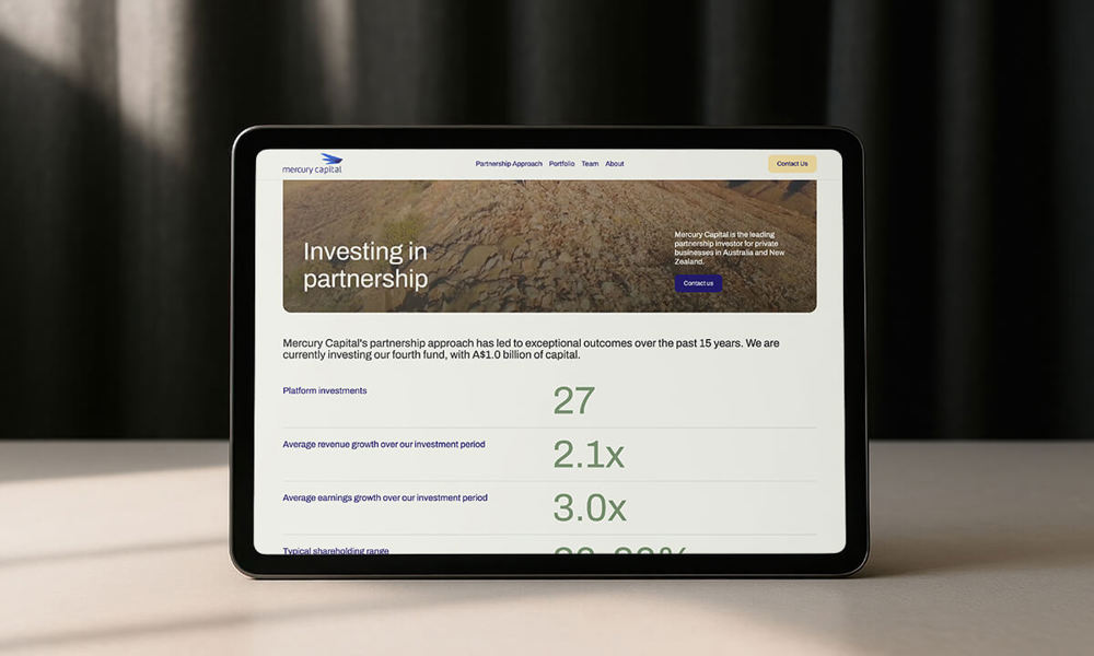 A screenshot of the new Mercury Capital homepage, shown on a tablet computer on a pale wooden desk with dark grey curtains behind.