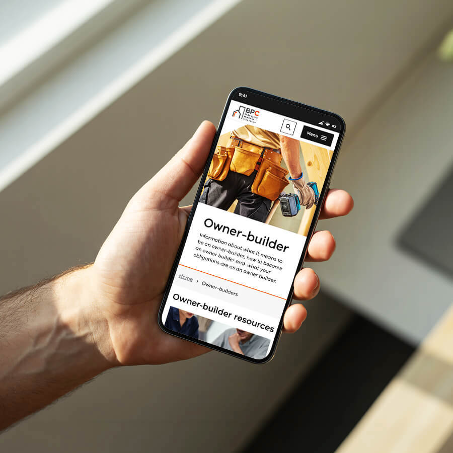 A man in his living room, standing by a window, holding his mobile phone that has the owner-builder webpage of the BPC website open on-screen.